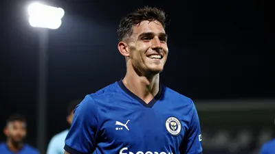 A young male footballer in a blue jersey, mid-20s, celebrating with a smile after a successful trial match